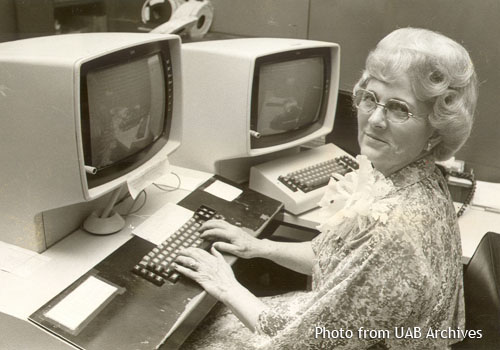 A woman sits in front of a computer and looks to the side at the camera