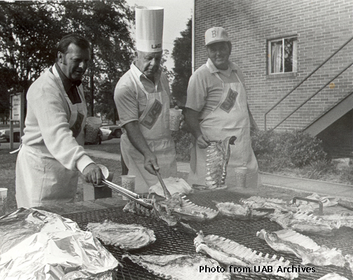 Three men in aprons tend to a bbq pit